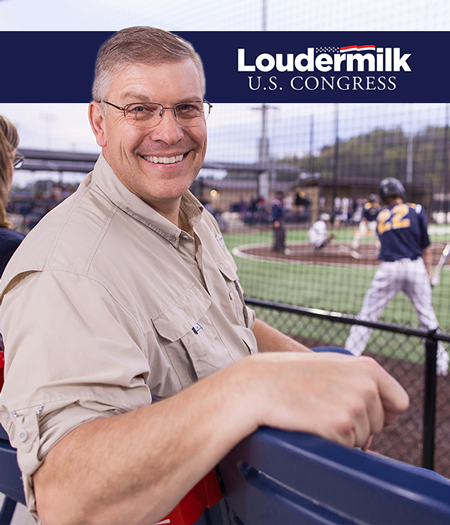 Barry-loudermilk-mid-50s-caucasian-male-sitting-on-bench-turnt-around-smiling-beyond-fence-of-baseball-game-wih-campaign-sign-on-top