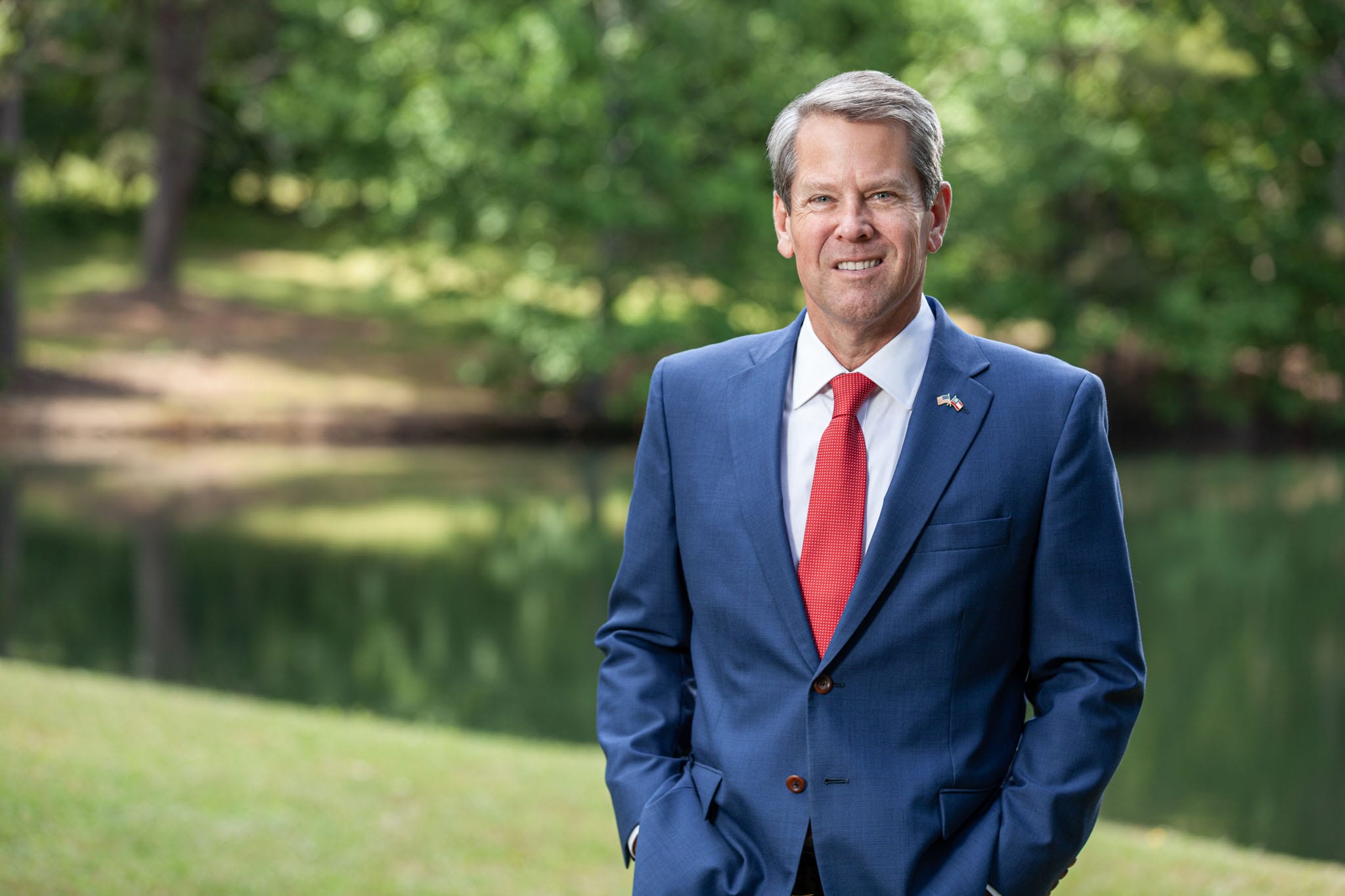 Georgia Governor Kemp standing in front of a pond in a blue suit with a red tie.